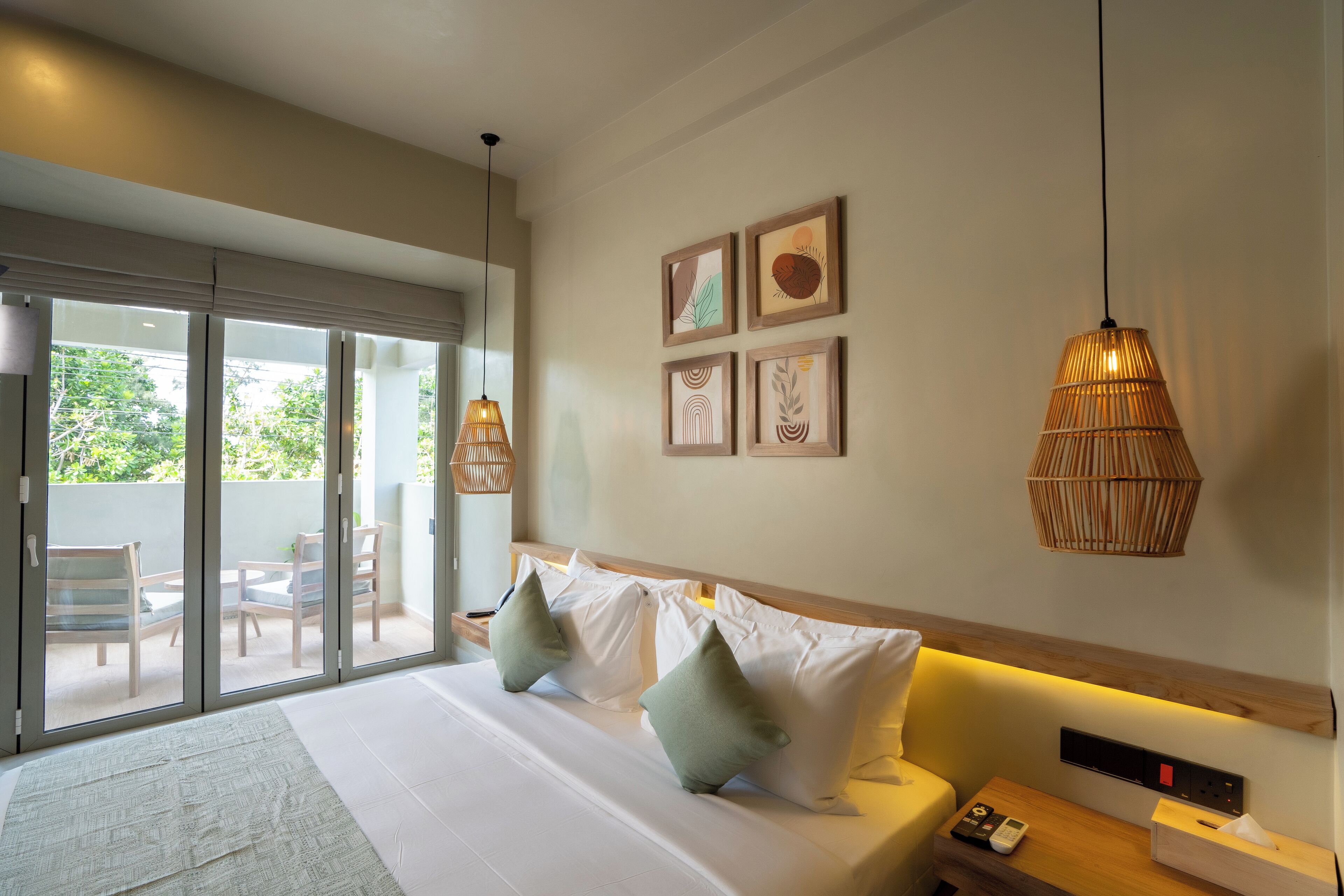 A modern hotel room with a large bed, a bedside table with a remote control, and a view of a balcony with outdoor furniture.