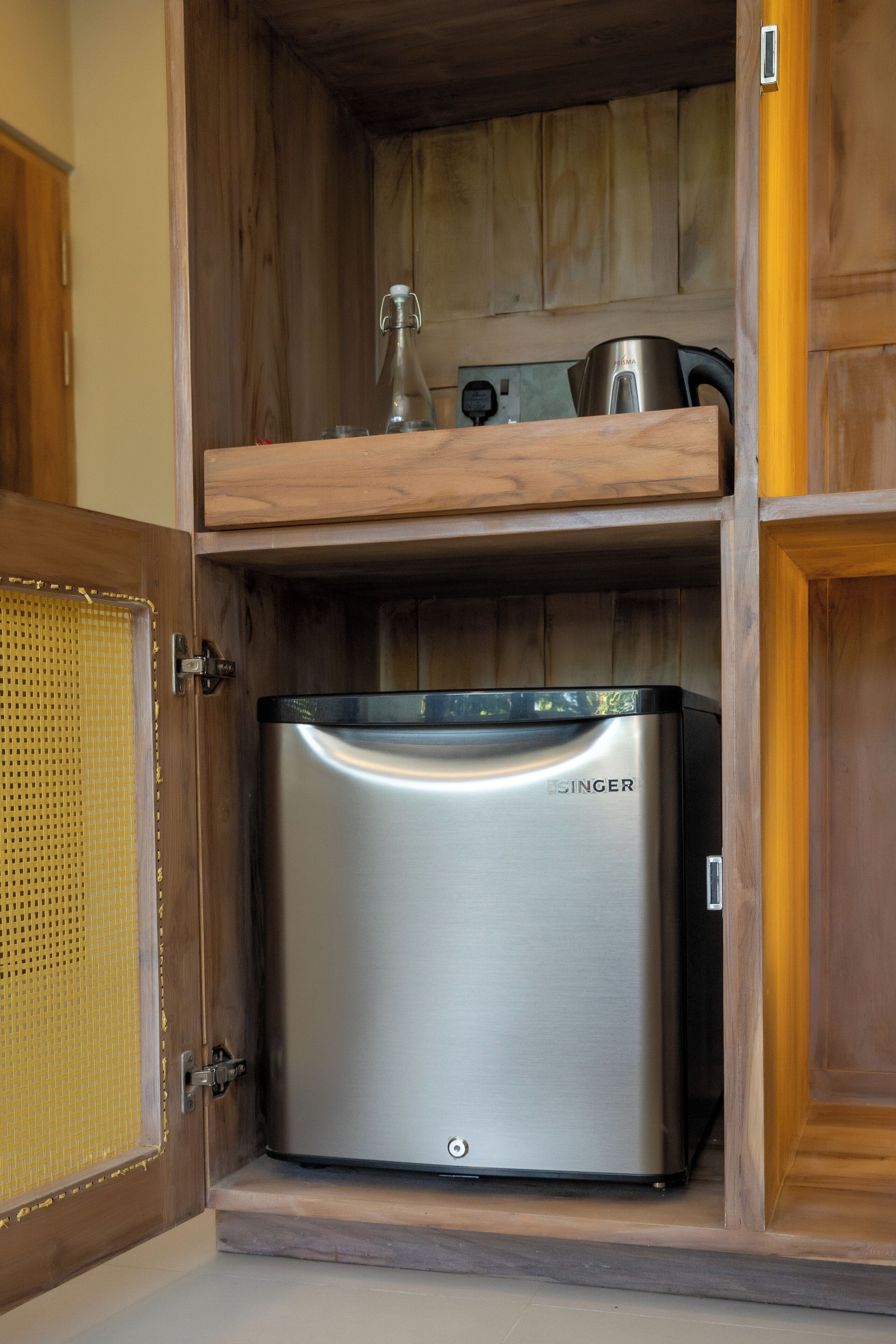 A compact kitchen with a stainless steel refrigerator, a wooden shelf above, and a woven basket door.