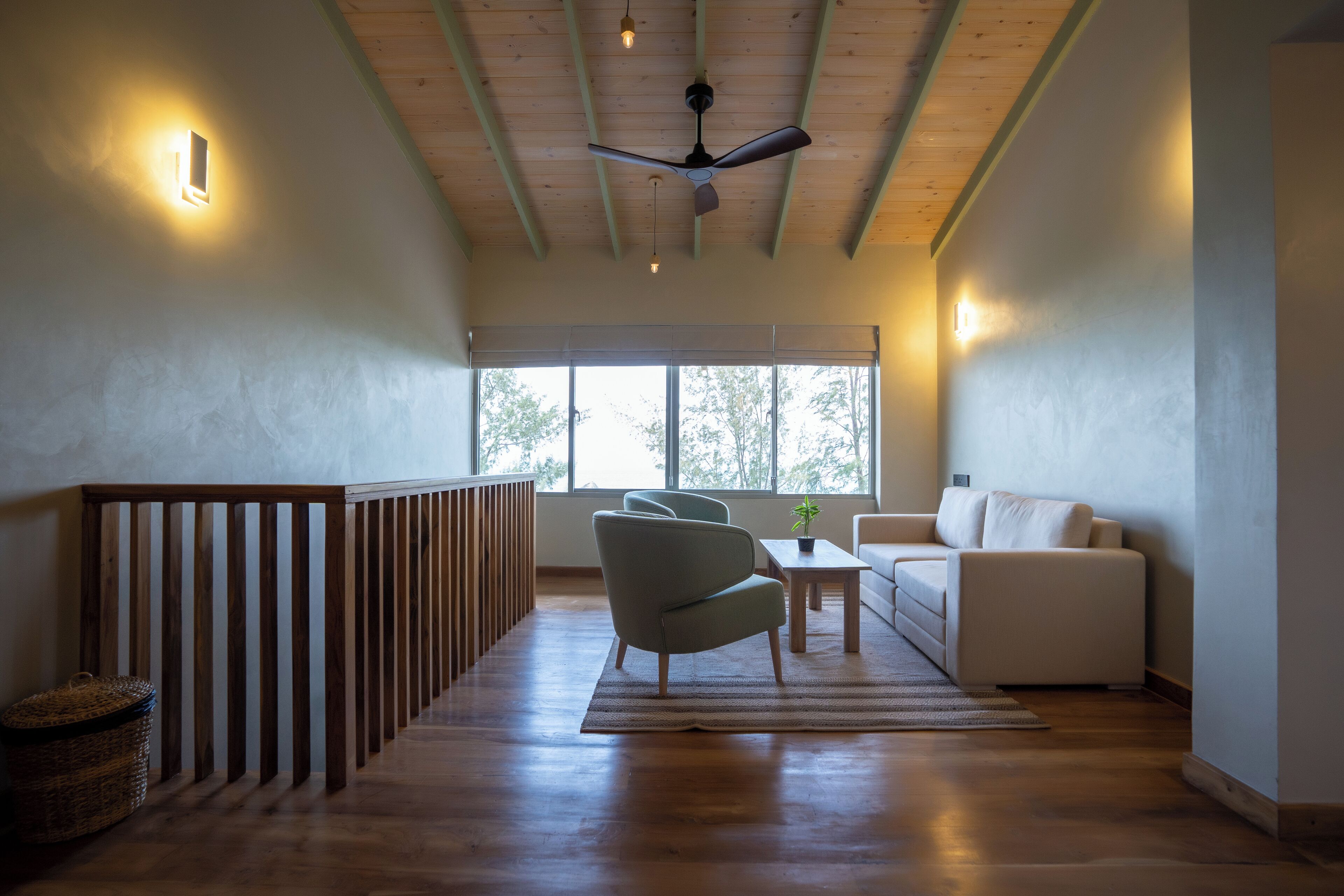 A spacious room with a wooden ceiling, a ceiling fan, a large window with a view of trees, a white sofa, and a light-colored armchair.