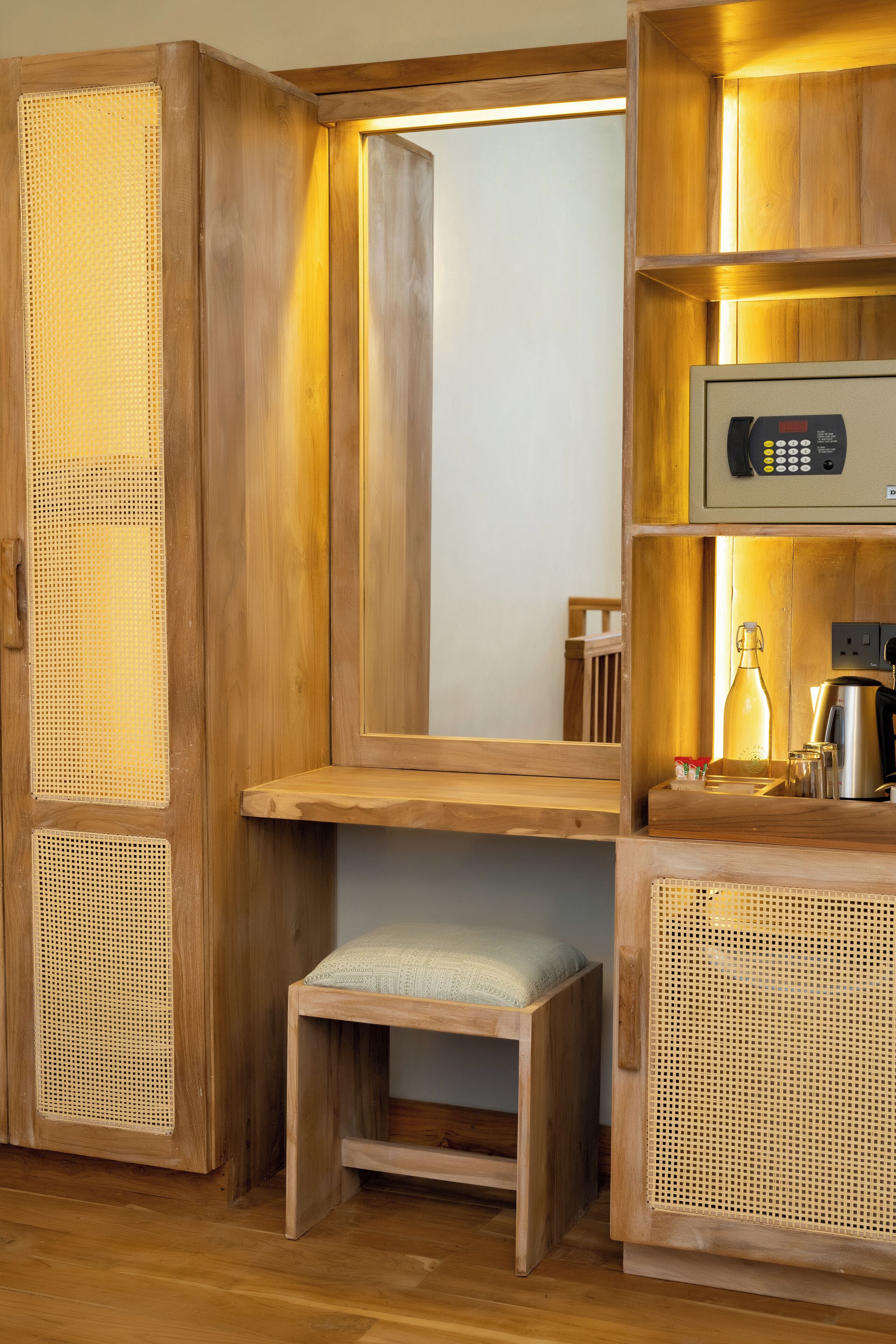 A wooden vanity with a mirror, a small stool, and a shelf with a coffee maker and a bottle.