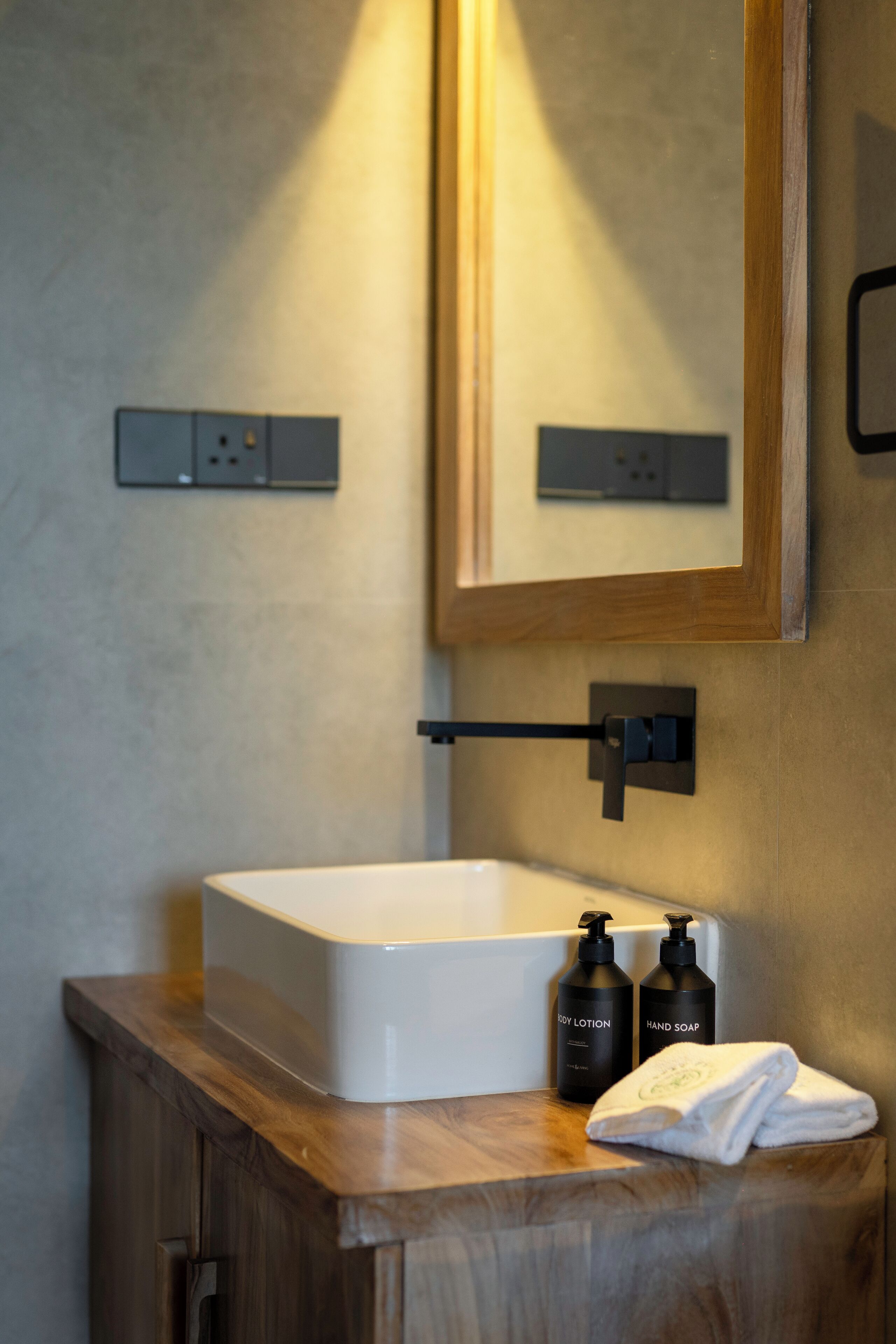 Ein Badezimmer mit einem weißen Waschbecken, zwei schwarzen Flaschen mit den Aufschriften 'Body Lotion' und 'Handseife' sowie einem Handtuch auf einer Holzauskład.