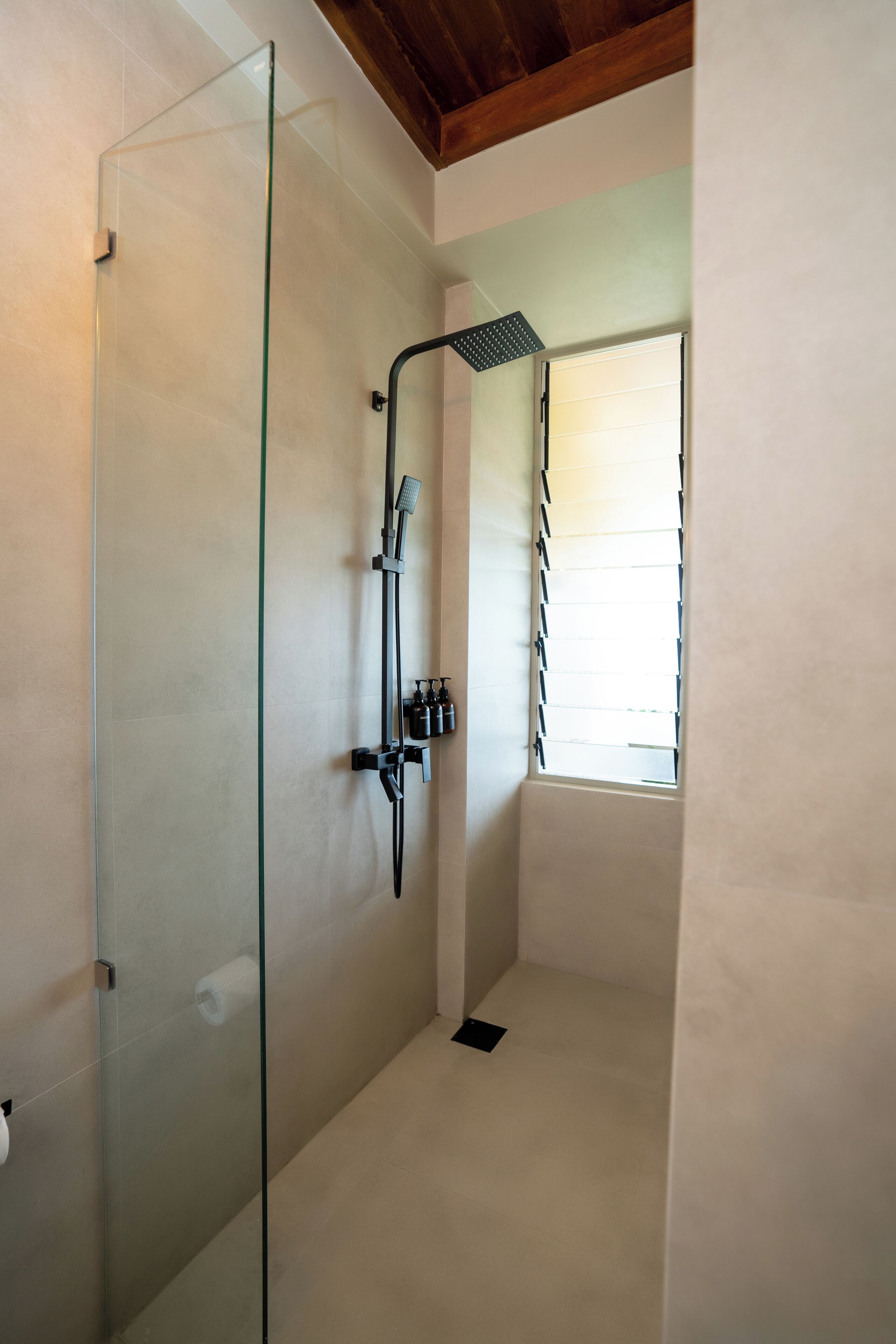A modern bathroom with a glass shower enclosure, a handheld showerhead, and a window with blinds.
