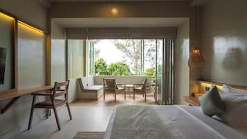 A modern hotel room with a large bed, a seating area with chairs and a table, and a balcony with a view of greenery.
