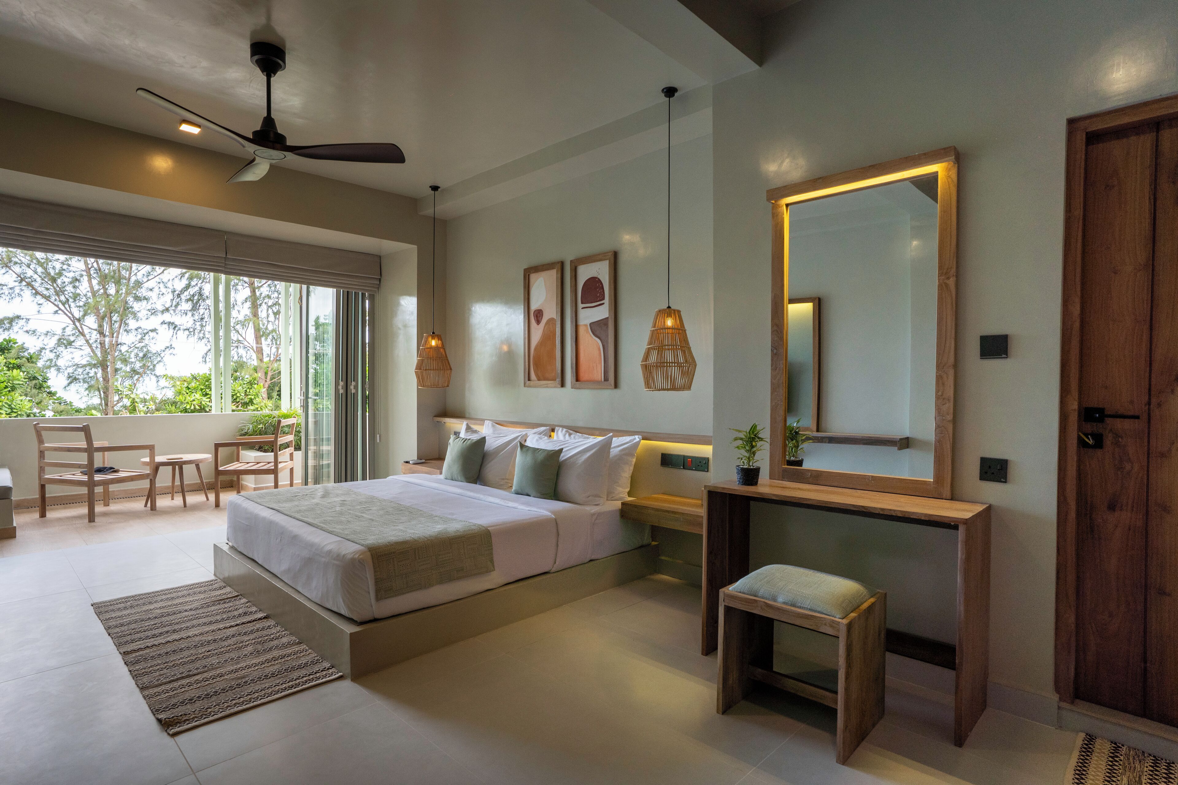 Une chambre moderne avec un grand lit, une table de chevet et un miroir. On y trouve un ventilateur de plafond, une fenêtre avec des stores et une vue sur les arbres à l’extérieur.