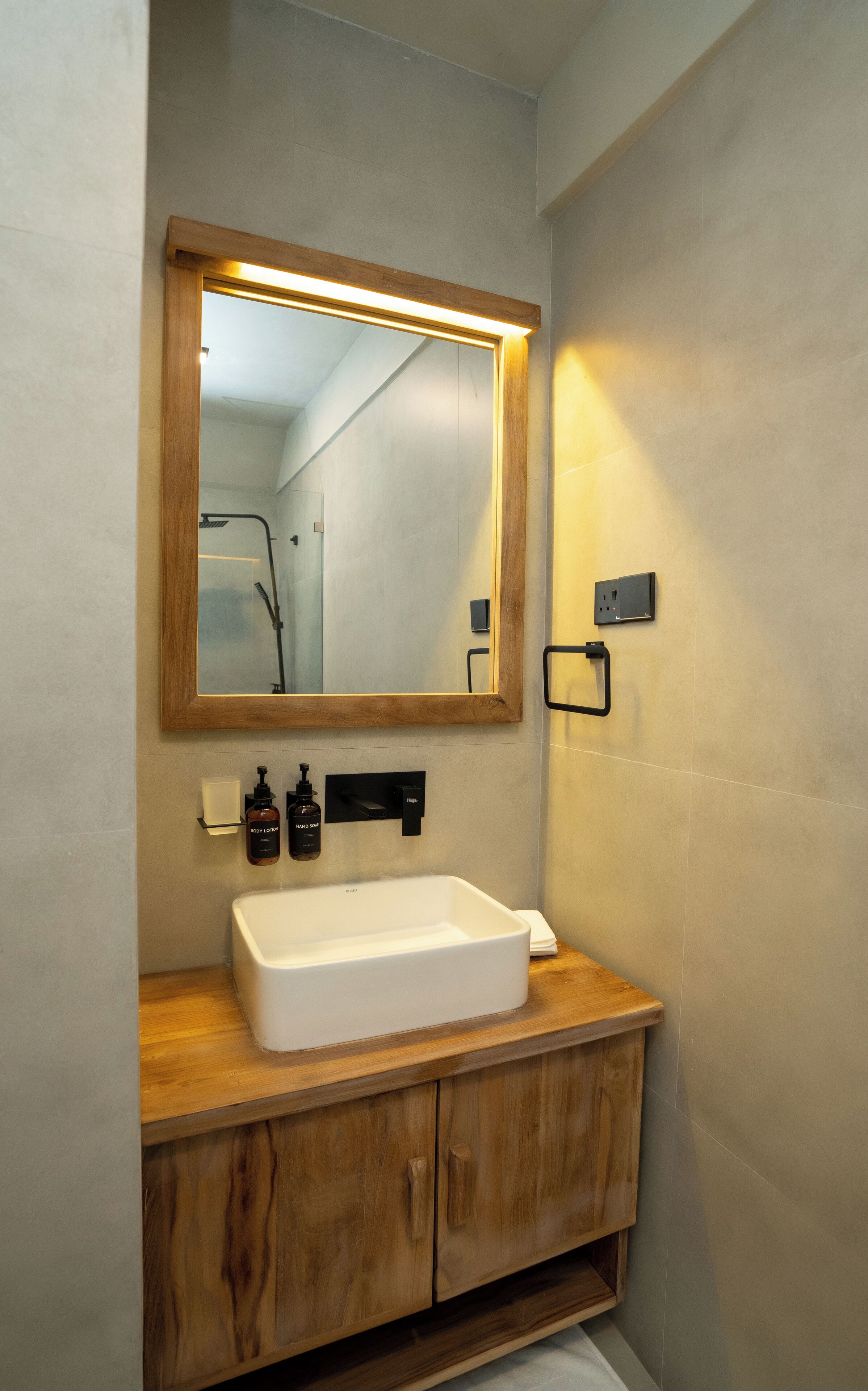 Een badkamer met een houten wastafelmeubel, een witte wastafel, een spiegel met houten lijst en een wandlamp.