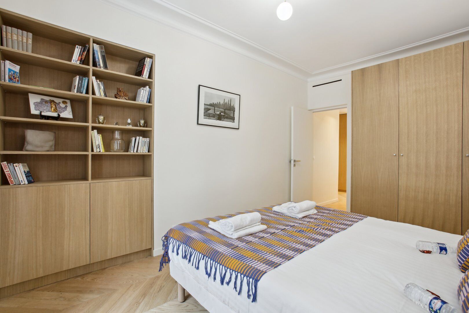 Een hotelkamer met een bed, een houten kledingkast, een boekenplank en een lijst aan de muur.
