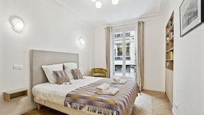 Un dormitorio moderno con una cama grande, una mesita de noche, una silla, una ventana con cortinas y una estantería.