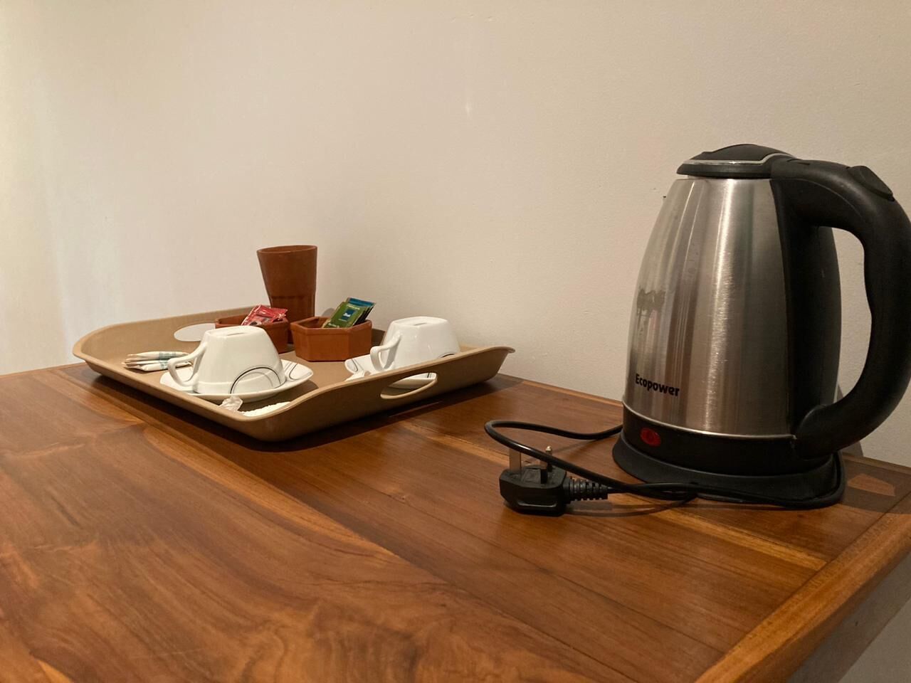 A wooden table with a tray holding a white cup, a small container, and a smartphone. Next to it is a stainless steel kettle with a black handle and a power cord.