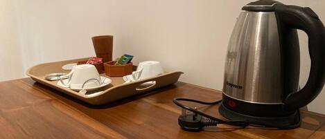 A wooden table with a tray holding a white cup, a small container, and a smartphone. Next to it is a stainless steel kettle with a black handle and a power cord.