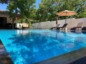 A clear swimming pool with lounge chairs and an umbrella, surrounded by a white wall and trees. - Kirinda Paradise Yala (Kirinda)