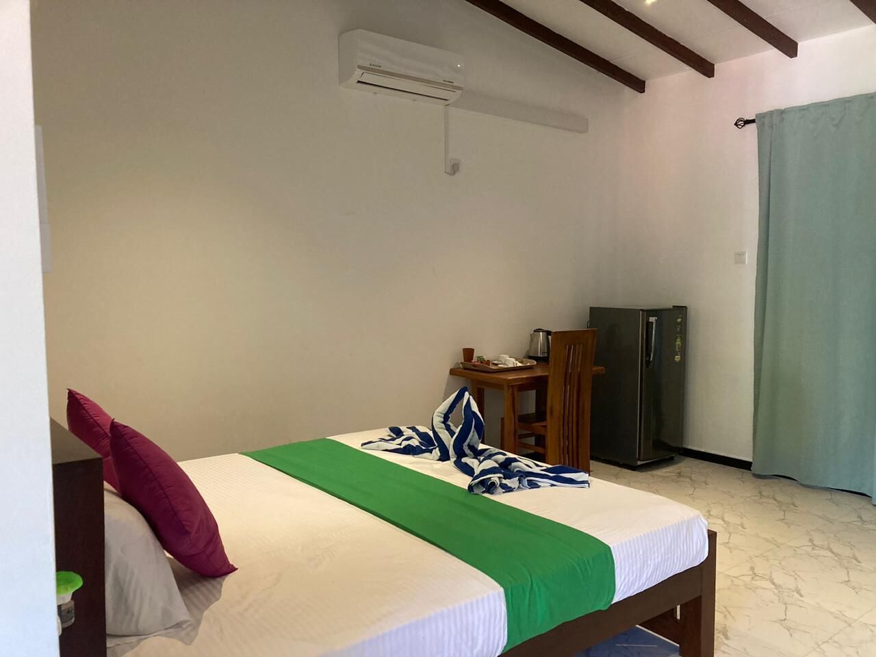 A single bed with a green and white bedspread, a wooden nightstand with a kettle, and a refrigerator in a room with white walls and a marble floor.