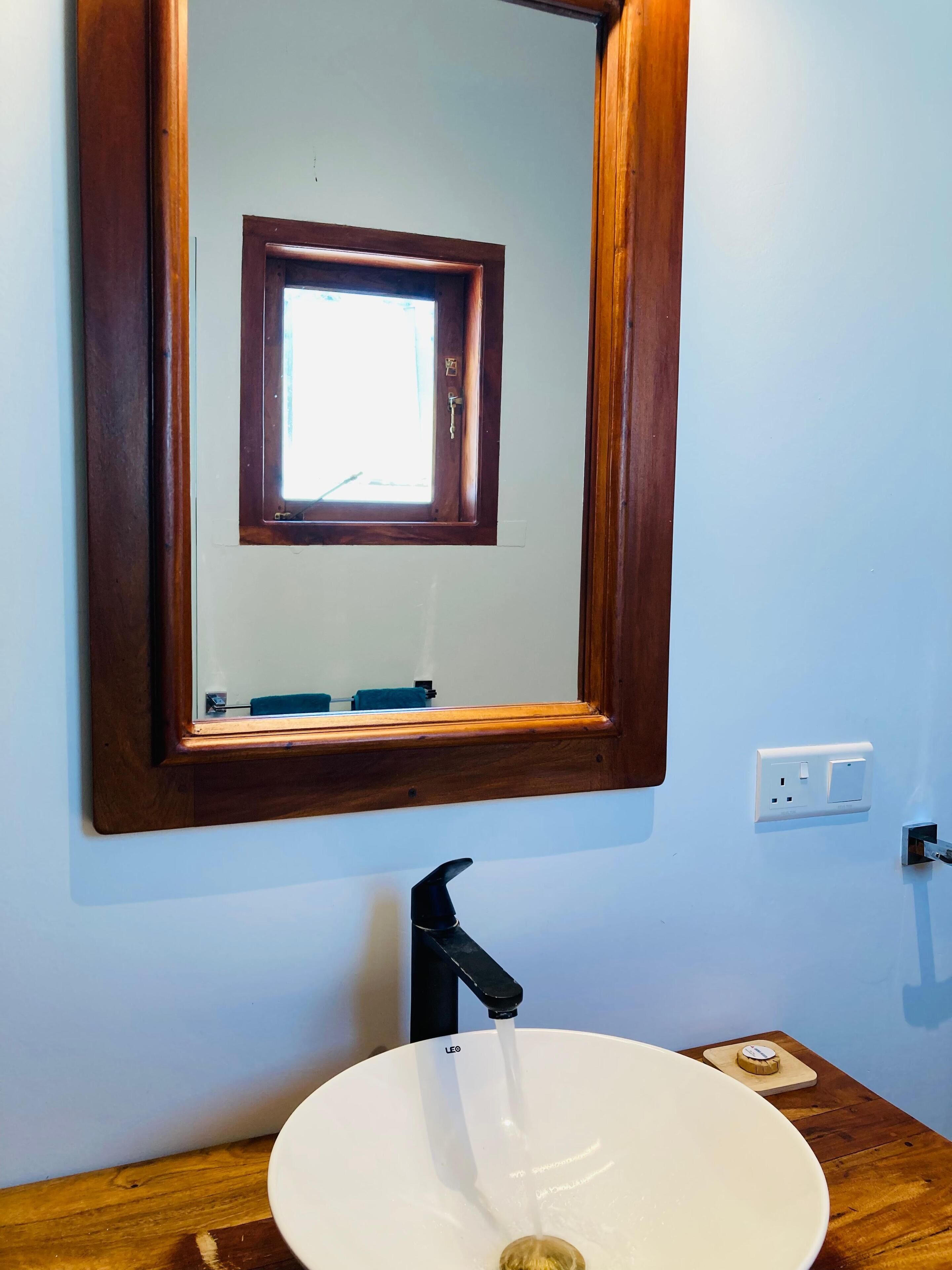 Une salle de bain avec un miroir encadré en bois, un lavabo blanc et une fenêtre située au-dessus du miroir.