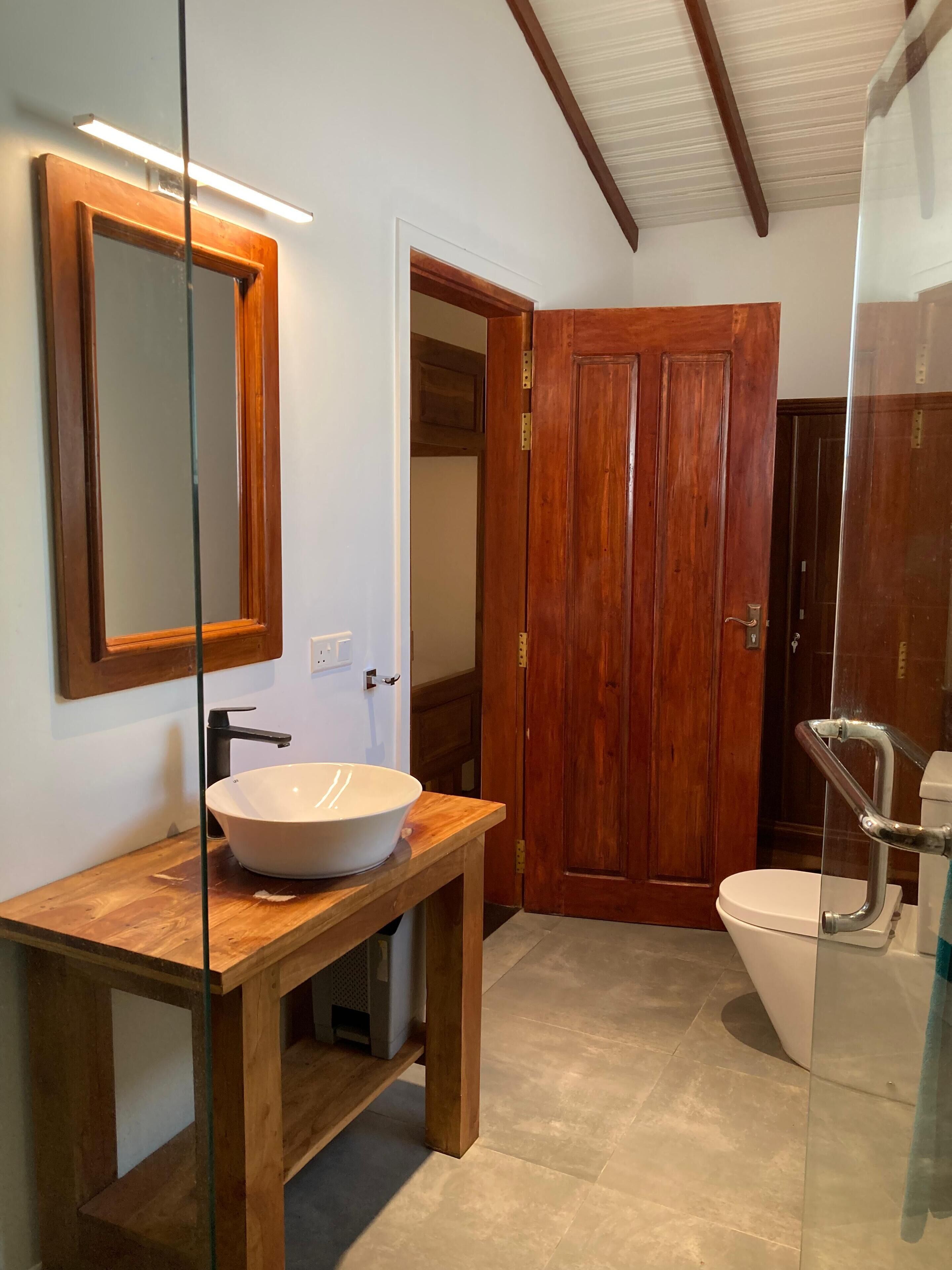 Ein Badezimmer mit einem Holzwaschtisch, einem großen Spiegel, einem weißen Waschbecken und einer Toilette mit Glasabtrennung.