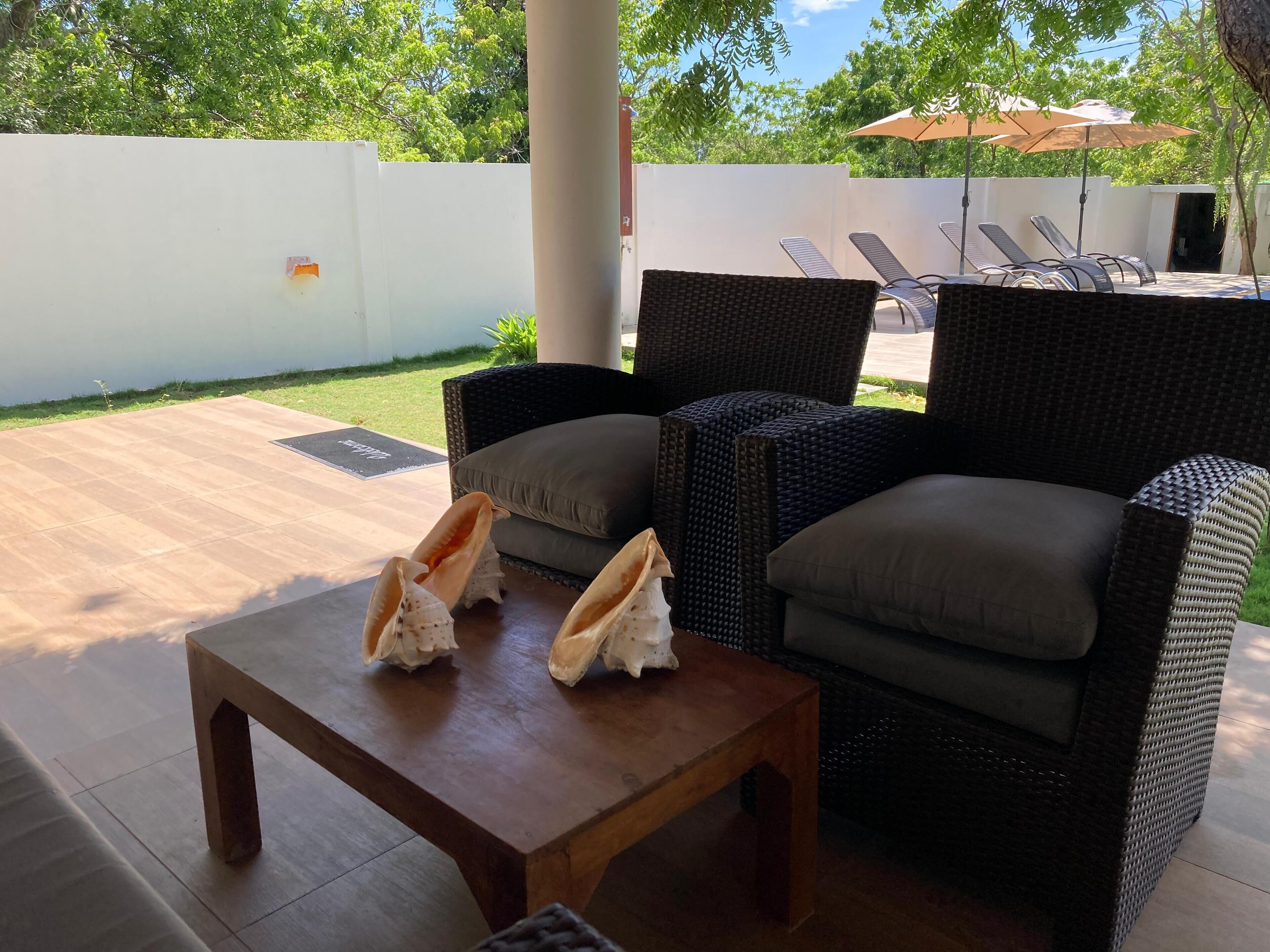 An outdoor seating area with wicker chairs and a wooden table, a white wall, and a view of a garden and trees in the background.
