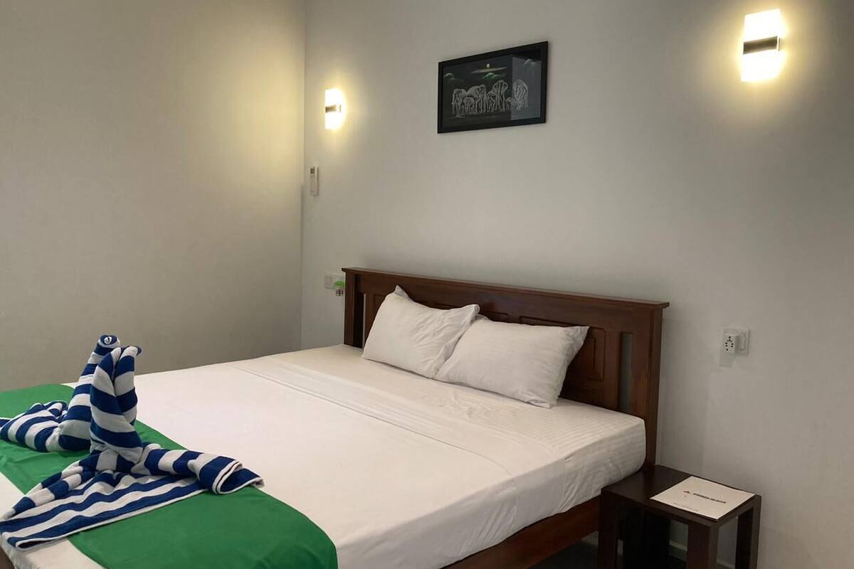 Un lit bien fait, avec du linge de lit blanc et une serviette à rayures vertes et bleues. Il y a une petite table de chevet avec une lampe, et un tableau encadré est accroché au mur au-dessus du lit.