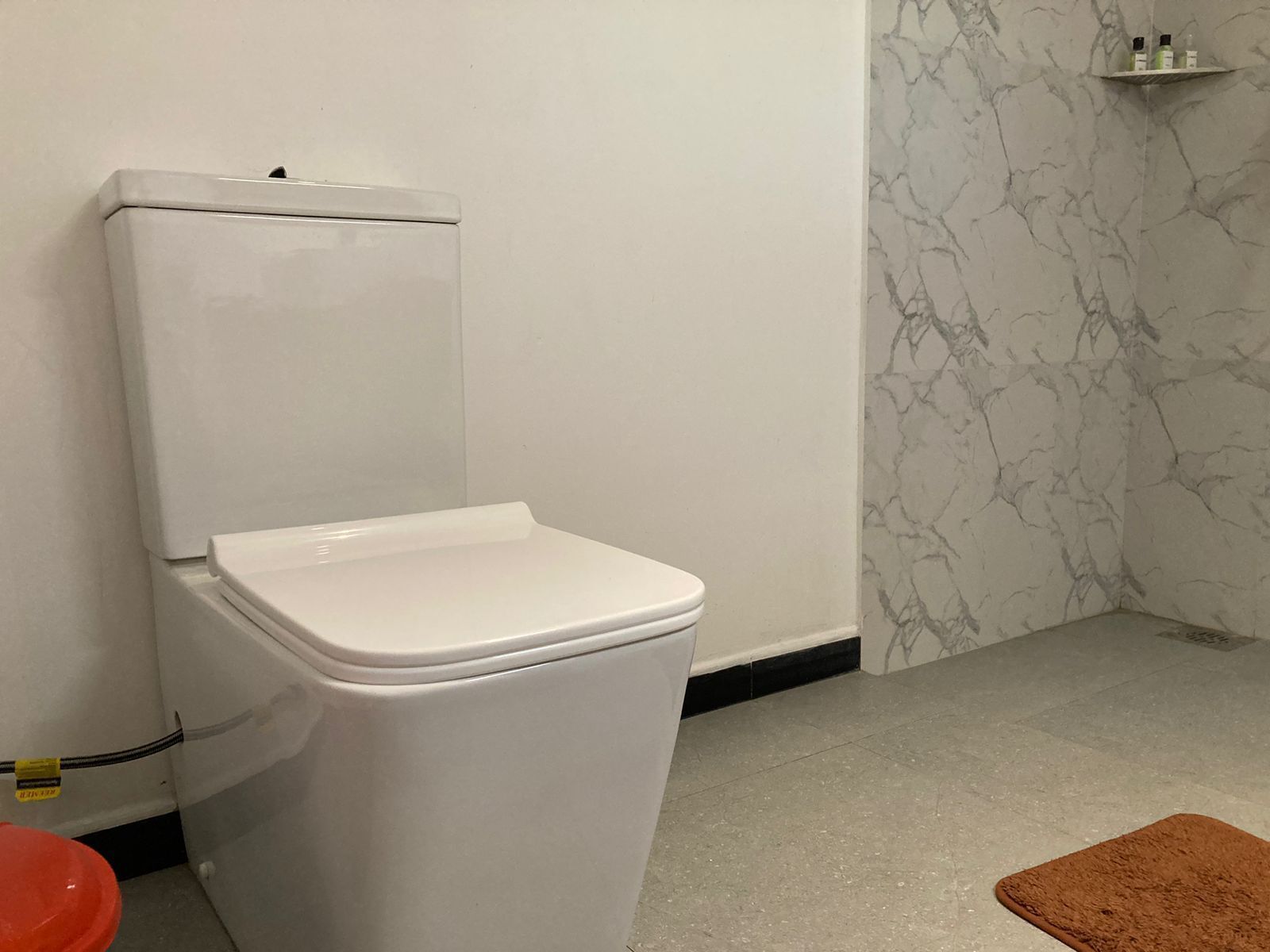 Des toilettes blanches dans une salle de bain avec un mur en marbre et une petite étagère contenant des articles de toilette.