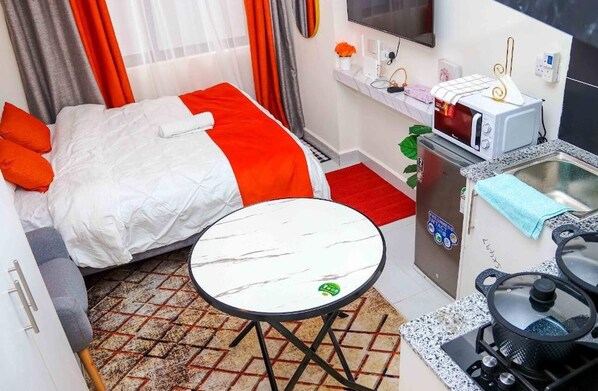 Una habitación de hotel pequeña y funcional con cama, una mesita redonda, una kitchenette con microondas y lavabo.