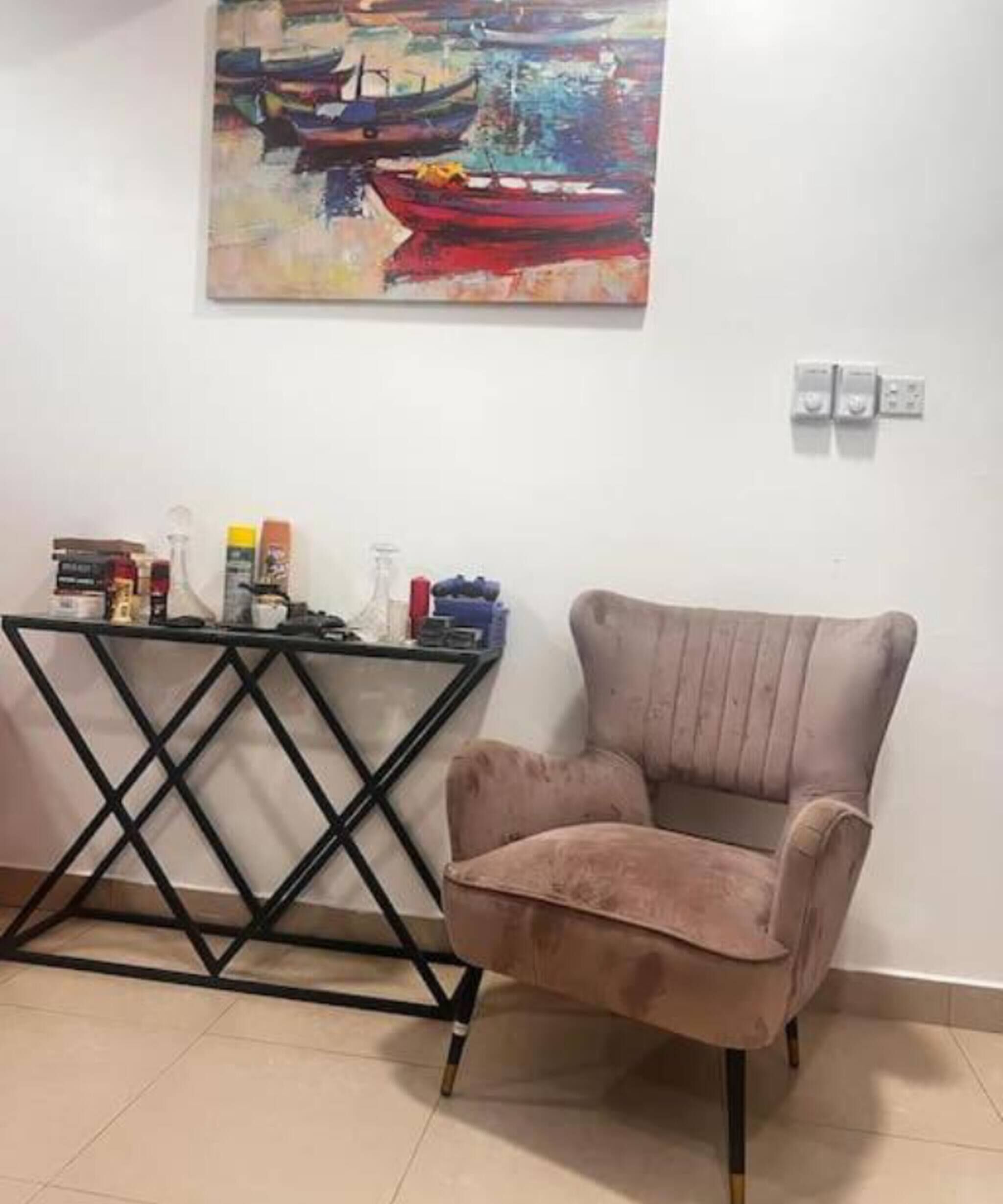 Una habitación con un sillón color beige, una mesita consola de metal negro con objetos encima, y un cuadro de barcos en la pared.