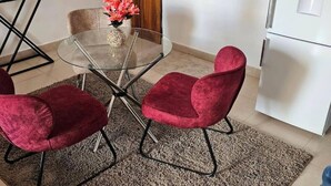Un comedor con una mesa de vidrio, dos sillas rojas, un florero con flores y una cocina al fondo.