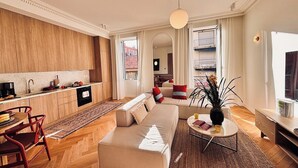 A modern living room with a sofa, coffee table, and dining area. There is a kitchen in the background with wooden cabinets. - Luxury Apartment - 2 bedrooms (Nice)