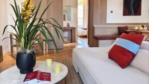 A modern hotel room with a round coffee table, a potted plant, a sofa with a red and blue pillow, and a bed in the background. - Luxury Apartment - 2 bedrooms (Nice)