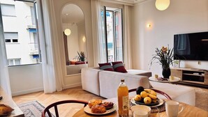A modern living room with a dining area, featuring a sofa, a TV, and a breakfast setup with croissants and fruit. - Luxury Apartment - 2 bedrooms (Nice)