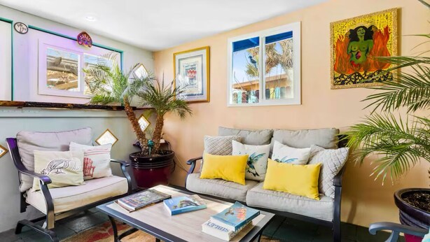 Un salon avec un canapé, des fauteuils et une table basse sur laquelle sont posés des livres. On y trouve également des plantes en pot et des œuvres d’art accrochées aux murs.