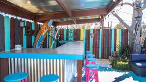Un espace bar extérieur abrité, doté d’une clôture en bois, de tabourets colorés et d’un comptoir en métal.