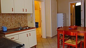 A compact kitchen with white cabinets, a sink, and a refrigerator. There is a dining table with chairs and a bowl on it. - Gallipoli Ubicación Chiesanuova (Sannicola)