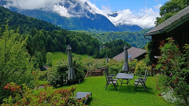 A lush green garden with a patio area, a mountain backdrop, and a traditional building. - Holiday Home 'Ferienhaus-Bregenzerwald' with Mountain View, Private Terrace and Wi-Fi (Bizau)