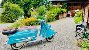A vintage blue scooter parked on a gravel path, a wooden house with a sloped roof, and a bench with a table nearby. - Holiday Home 'Ferienhaus-Bregenzerwald' with Mountain View, Private Terrace and Wi-Fi (Bizau)