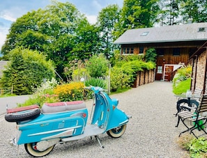 A vintage blue scooter parked on a gravel path, a wooden house with a sloped roof, and a bench with a table nearby.