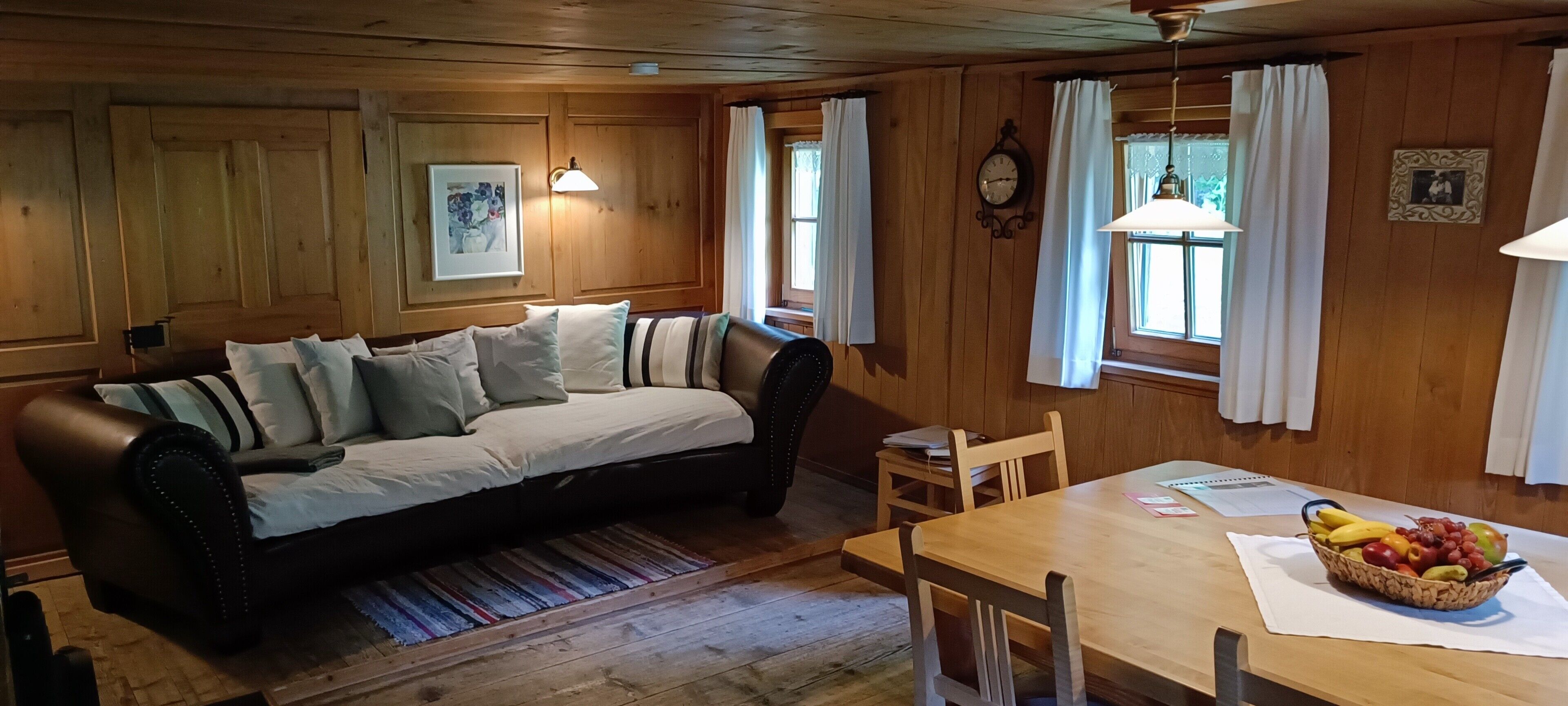 Ein gemütliches Zimmer mit Holzboden, einer Couch mit Kissen, einem Esstisch mit Stühlen, einer Uhr und einer Obstschale.