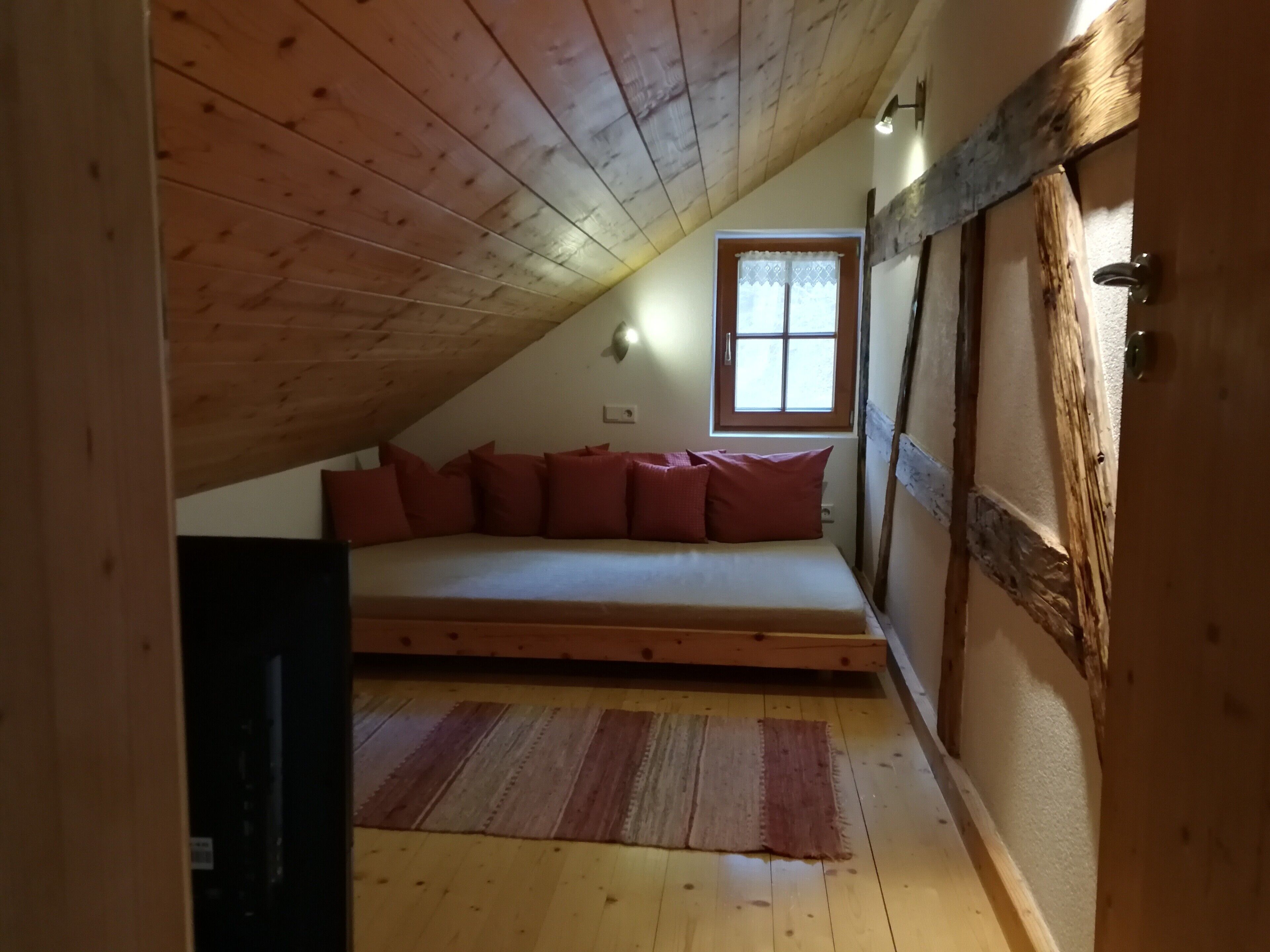 Un dormitorio tipo loft con techo inclinado, una cama individual con almohadas rojas, una ventana con cortinas de encaje y una puerta de madera.