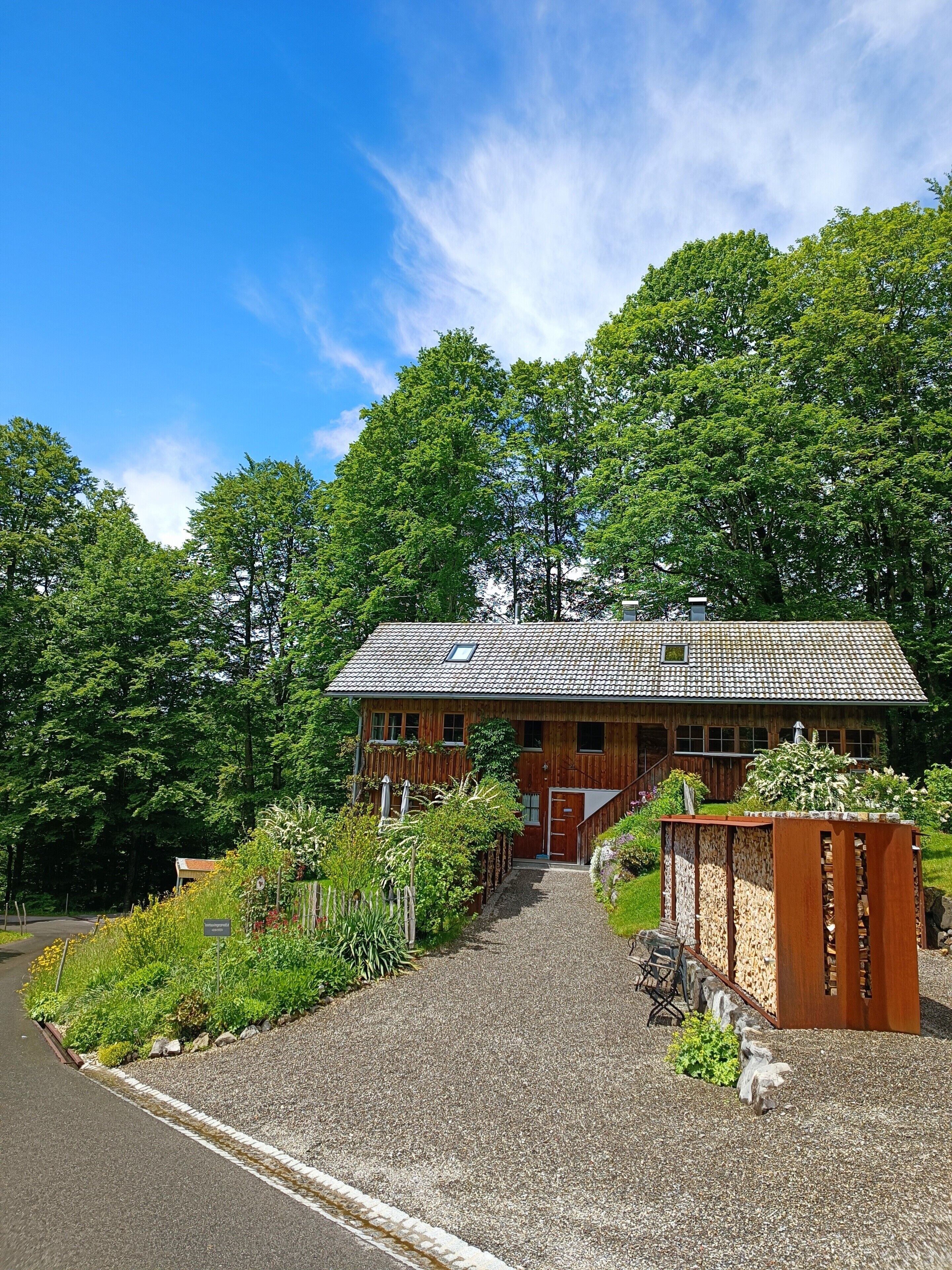 A wooden house with a gravel driveway, surrounded by lush greenery and trees.