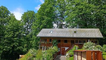 A wooden house with a gravel driveway, surrounded by lush greenery and trees.