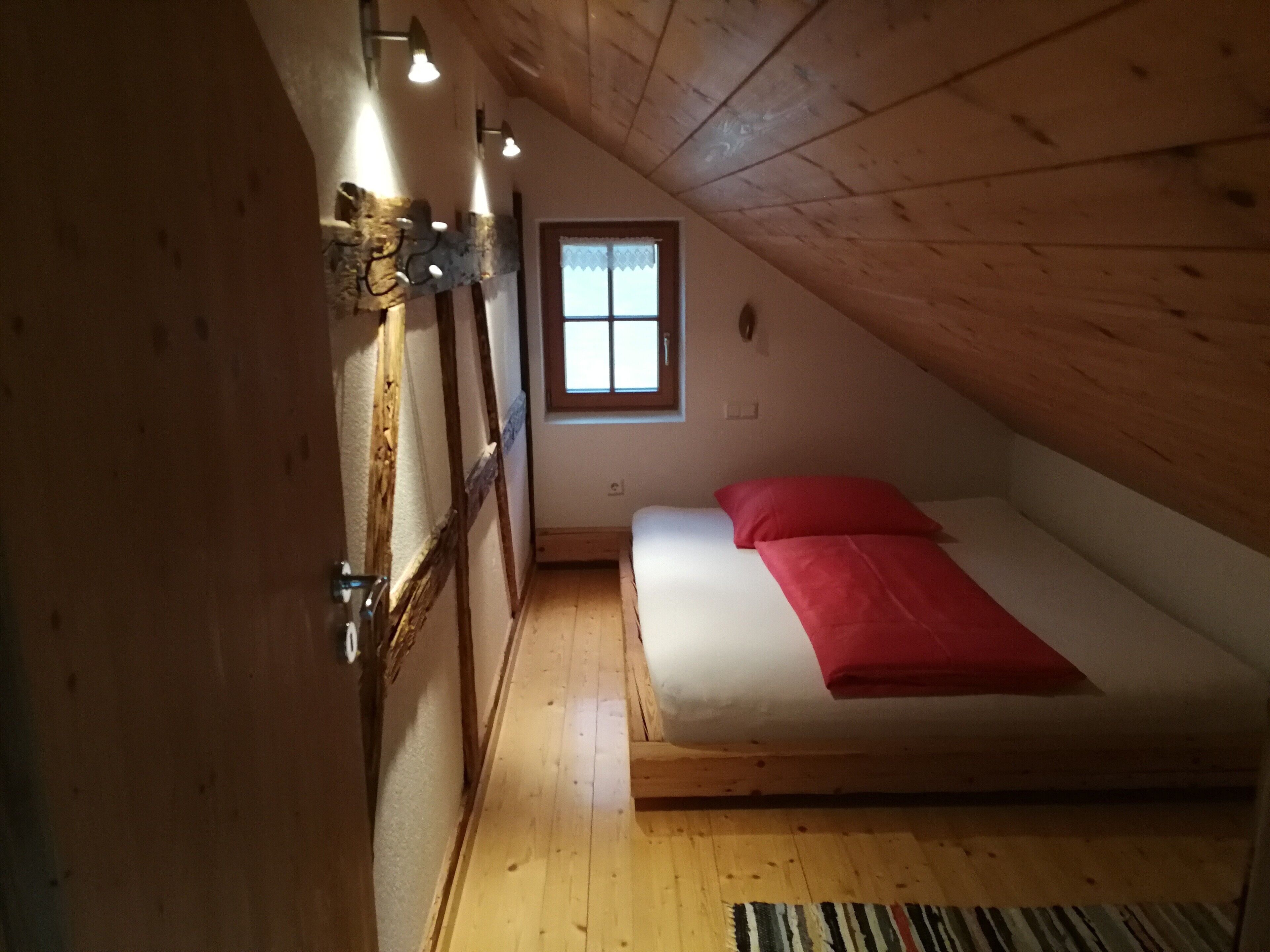 Une petite chambre mansardée, chaleureuse et accueillante, avec un plafond en bois, un lit simple avec une literie rouge, une fenêtre ornée de rideaux blancs et un plancher en bois.