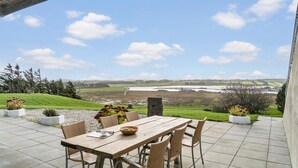 Un patio exterior con mesa y sillas de madera, rodeado de plantas en macetas, con vistas a un paisaje pintoresco que incluye un río y colinas al fondo.