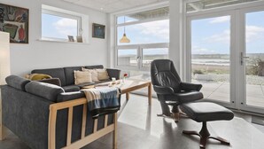 Una sala moderna con un sofá gris, un banco de madera, un sillón de cuero negro y una mesa de comedor. Amplias ventanas ofrecen vistas al exterior.