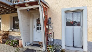 Een huis met een witte deur, een klein voortuintje, een metalen plankje met decoratieve spullen, en een raam met een witte zonwering.