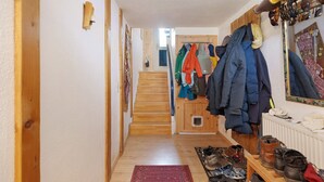 Een gang met houten vloer, een rood tapijt, een schoenenrek en een kapstok met verschillende jassen.