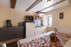 Une cuisine compacte équipée d’un four à micro-ondes, d’une machine à café et d’un petit réfrigérateur. On y trouve une table à manger avec une nappe à motifs et un panier. La pièce est dotée d’un plafond rustique avec des poutres apparentes et d’une fenêtre qui laisse entrer la lumière naturelle.