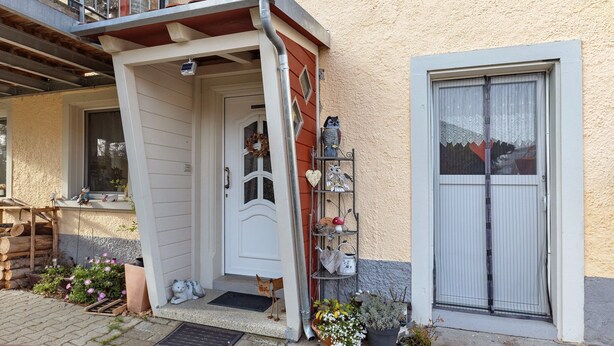 Una casa con puerta blanca, un pequeño porche, un estante metálico con adornos y una ventana con persiana blanca.