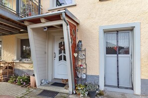 Una casa con puerta blanca, un pequeño porche, un estante metálico con adornos y una ventana con persiana blanca.