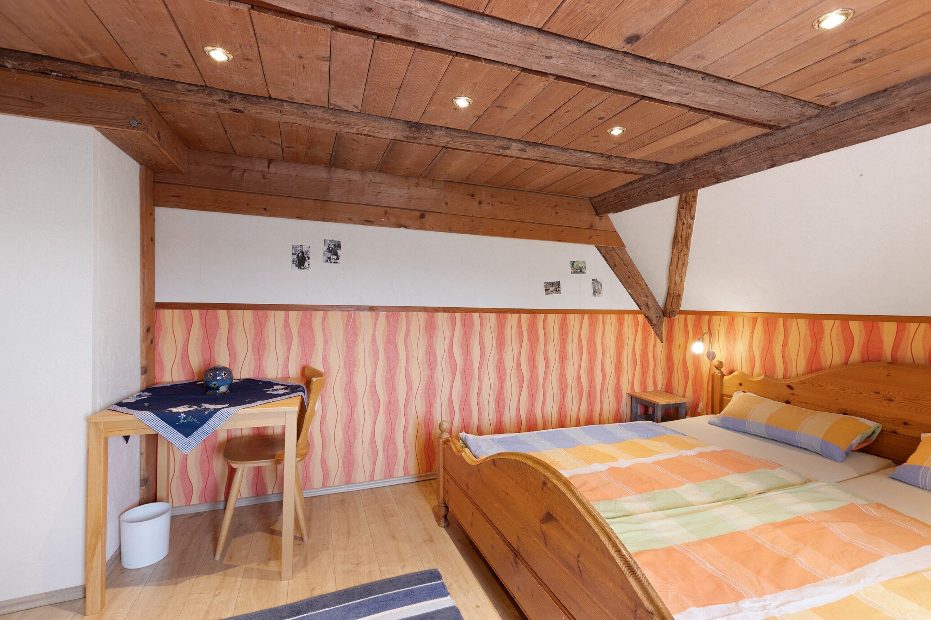 Une chambre avec un plafond en bois, un lit recouvert d’une courtepointe aux couleurs vives, un bureau avec une chaise et un mur orné d’un motif ondulé.