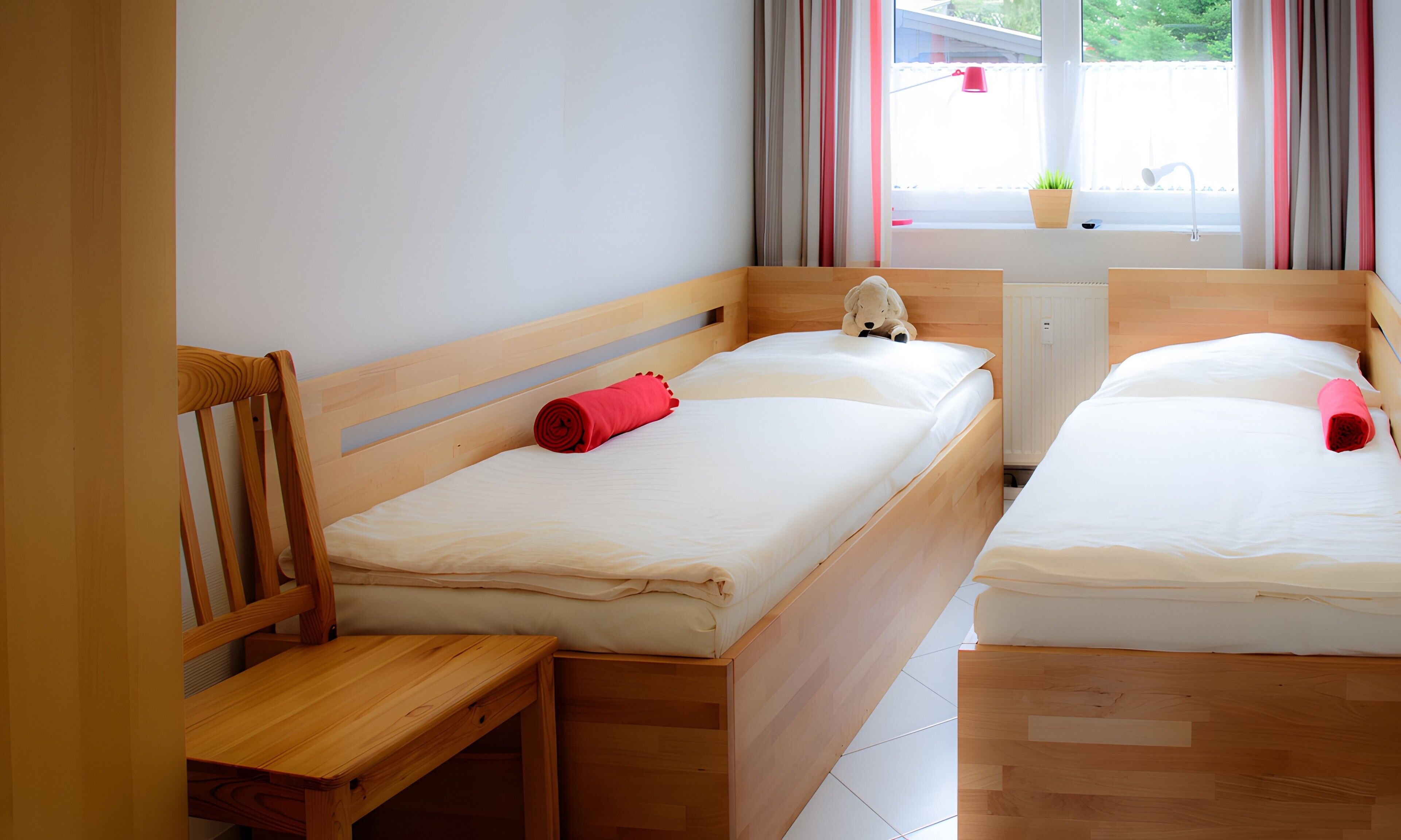 Um quarto com duas camas de madeira, uma cadeira de madeira, uma janela com cortinas e um radiador.