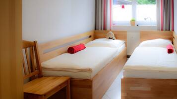 Um quarto com duas camas de madeira, uma cadeira de madeira, uma janela com cortinas e um radiador.