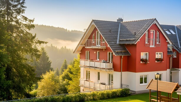 A red and white two-story house with a balcony, surrounded by lush greenery and a forested mountain backdrop. - Apartment 'Ferienwohnung Tannenblick 3-4' with Mountain View, Balcony and Wi-Fi (Oberhof)