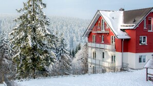 A red and white house with a balcony, surrounded by snow-covered trees and a fence. - Apartment 'Ferienwohnung Tannenblick 3-4' with Mountain View, Balcony and Wi-Fi (Oberhof)