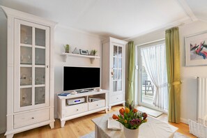 A living room with a television, white cabinets, a table with a vase of flowers, and a window with curtains. - Apartment 'Ferienwohnung Neumann' with Shared Garden, Balcony and Wi-Fi (Wesselburen)